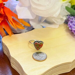 May include: A silver ring with a heart-shaped opal stone. The opal is a mix of red, orange, and green colors. The ring is next to a US dime for size comparison.