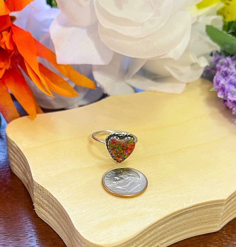 May include: A silver ring with a heart-shaped opal stone. The opal is a mix of red, orange, and green colors. The ring is next to a US dime for size comparison.