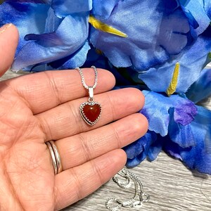 K&ouml;nnte beinhalten: Eine silberne Kettenkette mit einem herzf&ouml;rmigen Anh&auml;nger mit einem roten Karneol-Stein.
