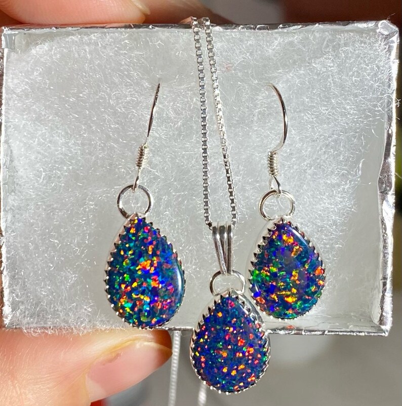 May include: A silver necklace and earrings set with teardrop-shaped black opal stones. The opals have a rainbow iridescent sheen.