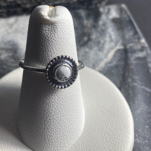 May include: A silver ring with a white howlite stone set in a decorative bezel. The ring is on a white ring display stand.