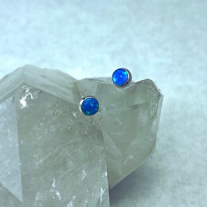 May include: Two small, round, blue opal earrings with silver posts. The earrings are sitting on a white crystal.