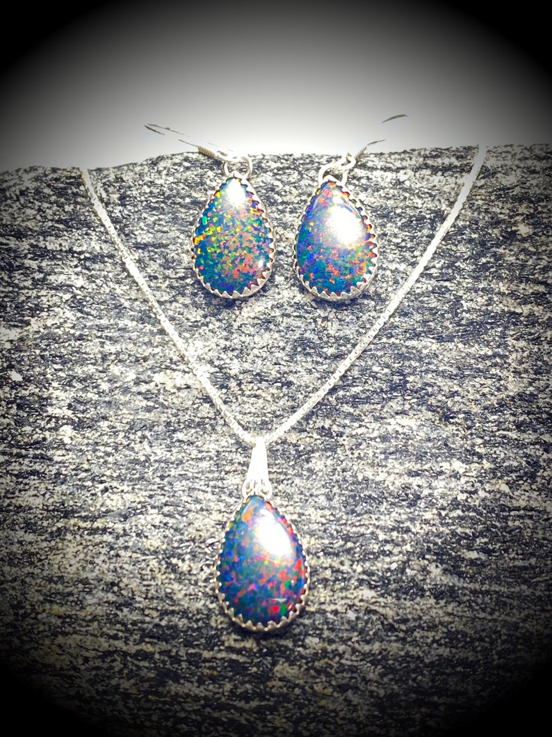May include: A silver necklace and earrings set with teardrop-shaped opal stones. The opals have a dark base with iridescent flashes of blue, green, and red.