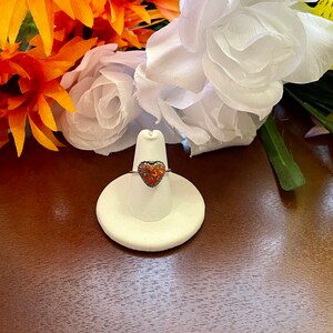 May include: A silver ring with a heart-shaped opal gemstone. The opal is a mix of red, orange, and yellow colors. The ring is displayed on a white ring holder.