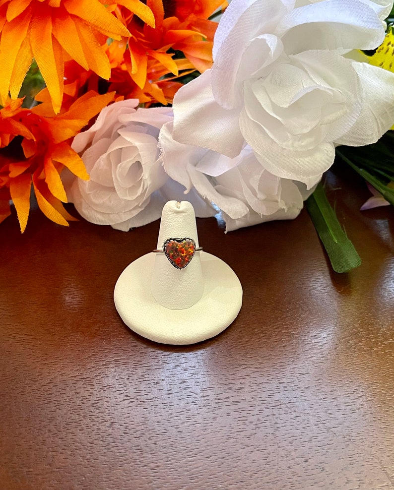 May include: A silver ring with a heart-shaped opal gemstone. The opal is a mix of red, orange, and yellow colors. The ring is displayed on a white ring holder.
