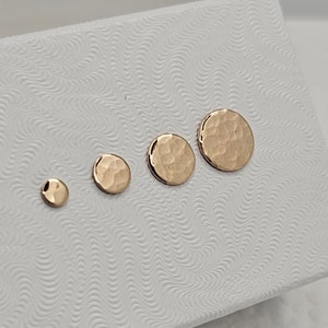 May include: Four gold-colored, hammered-finish stud earrings of varying sizes are displayed on a white textured surface. The earrings range from small, round studs to larger, circular designs. The earrings have a textured, hammered appearance.