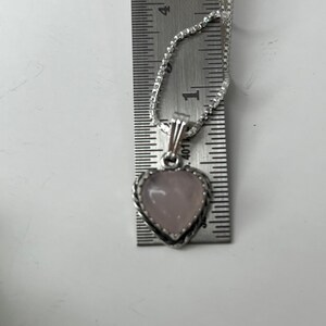 Puede incluir: Un collar de cadena de plata con un colgante en forma de coraz&oacute;n. El colgante est&aacute; hecho de una piedra rosa claro con un borde plateado. El colgante tiene un di&aacute;metro de aproximadamente 2,5 cm.