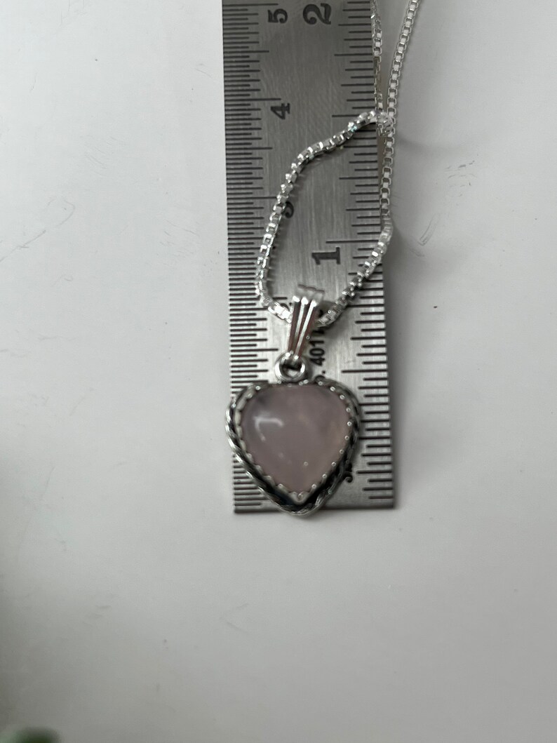 Puede incluir: Un collar de cadena de plata con un colgante en forma de coraz&oacute;n. El colgante est&aacute; hecho de una piedra rosa claro con un borde plateado. El colgante tiene un di&aacute;metro de aproximadamente 2,5 cm.