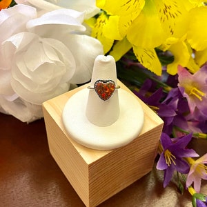 May include: A silver ring with a heart-shaped red opal gemstone. The ring is displayed on a white ring stand on a wooden surface.