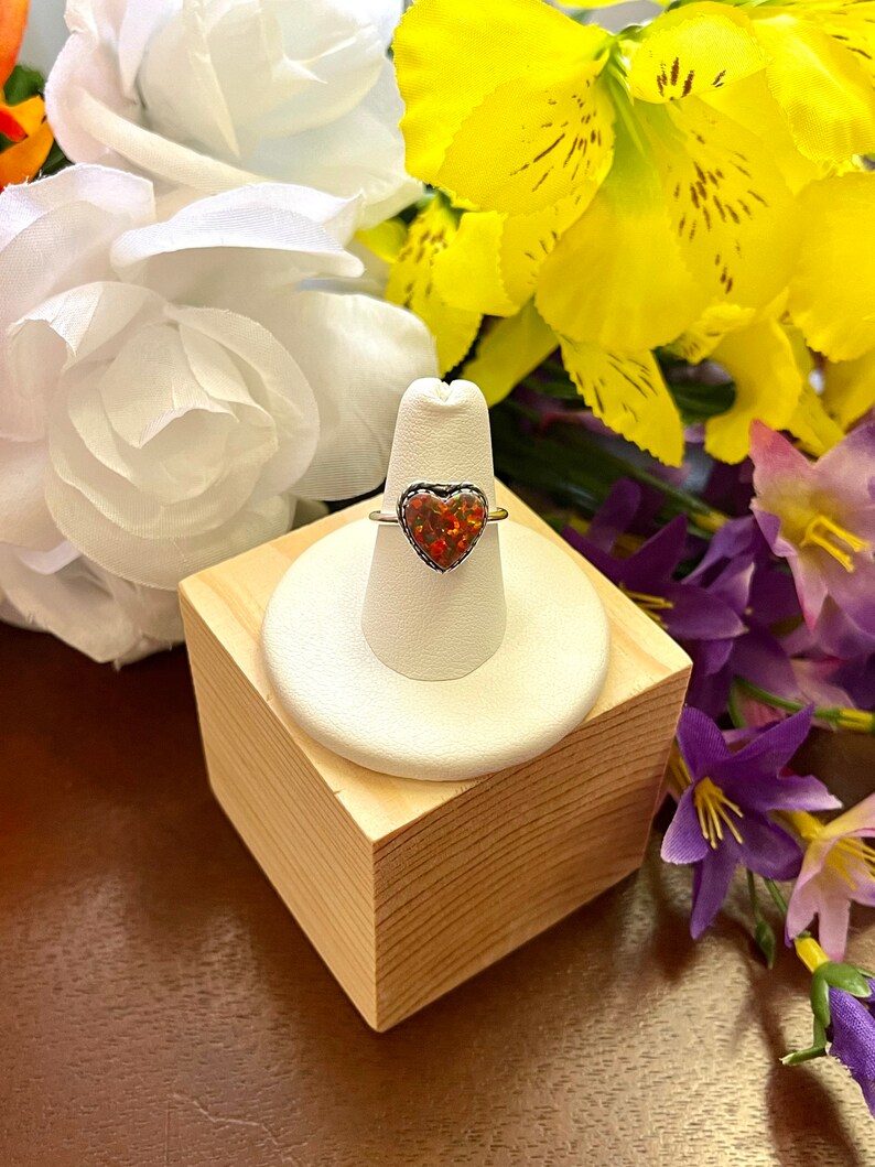 May include: A silver ring with a heart-shaped red opal gemstone. The ring is displayed on a white ring stand on a wooden surface.