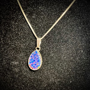 May include: A silver chain necklace with a teardrop-shaped pendant. The pendant is made of clear glass with a blue, pink, and white speckled design.
