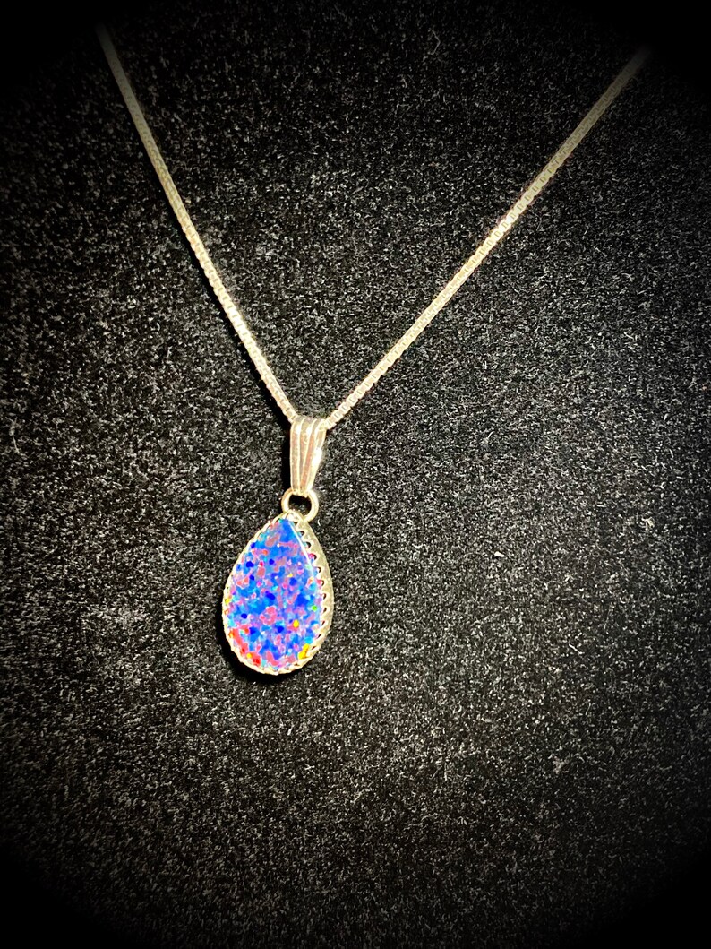 May include: A silver chain necklace with a teardrop-shaped pendant. The pendant is made of clear glass with a blue, pink, and white speckled design.
