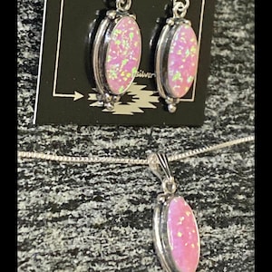 K&ouml;nnte beinhalten: Ein silbernes Halsketten- und Ohrring-Set mit rosafarbenen Opal-Steinen. Die Halskette hat eine zarte Kette und einen Anh&auml;nger mit einem ovalen rosafarbenen Opal-Stein. Die Ohrringe sind ebenfalls oval geformt mit rosafarbenen Opal-Steinen.