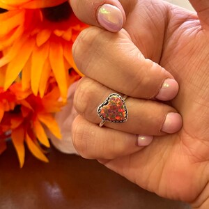 May include: A silver ring with a heart-shaped opal gemstone. The opal is a mix of pink, orange, and red.