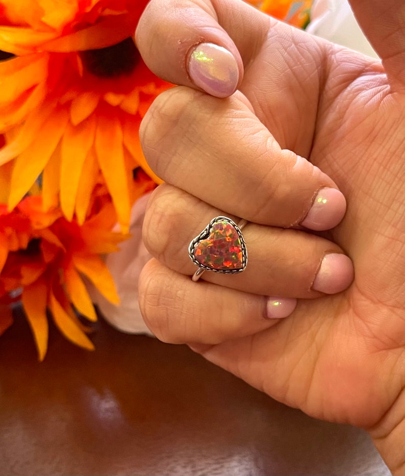 May include: A silver ring with a heart-shaped opal gemstone. The opal is a mix of pink, orange, and red.