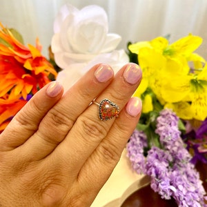 May include: A silver ring with a heart-shaped opal stone. The opal is a mix of pink, orange, and yellow.