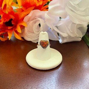 May include: A silver ring with a heart-shaped opal gemstone. The opal is a mix of orange, red, and yellow.