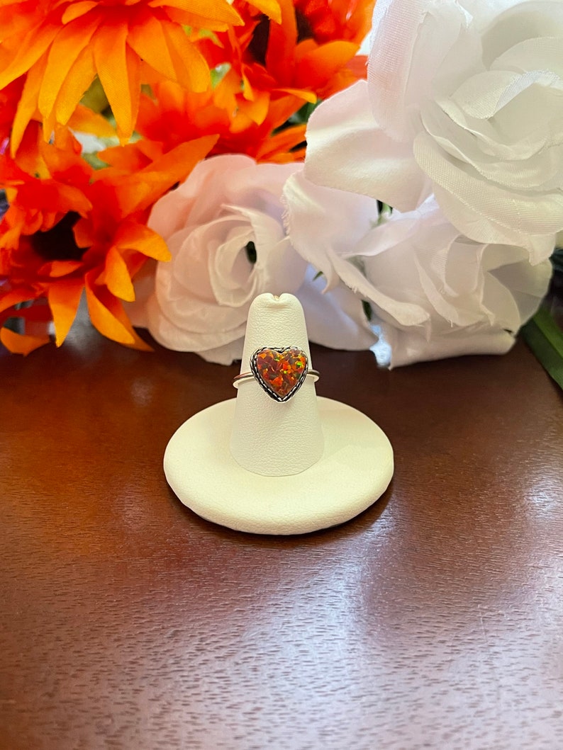 May include: A silver ring with a heart-shaped opal gemstone. The opal is a mix of orange, red, and yellow.