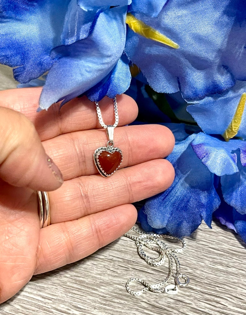 K&ouml;nnte beinhalten: Eine silberne Kettenkette mit einem herzf&ouml;rmigen Anh&auml;nger mit einem roten Karneol-Stein.