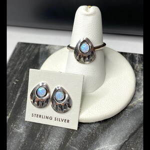 May include: A sterling silver ring and earrings set featuring a bear paw design with an iridescent opal stone in the center. The set is displayed on a white display stand.