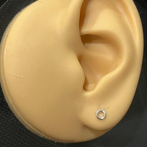 May include: A close-up of a silver hoop earring on a mannequin ear. The earring is a simple, thin hoop.
