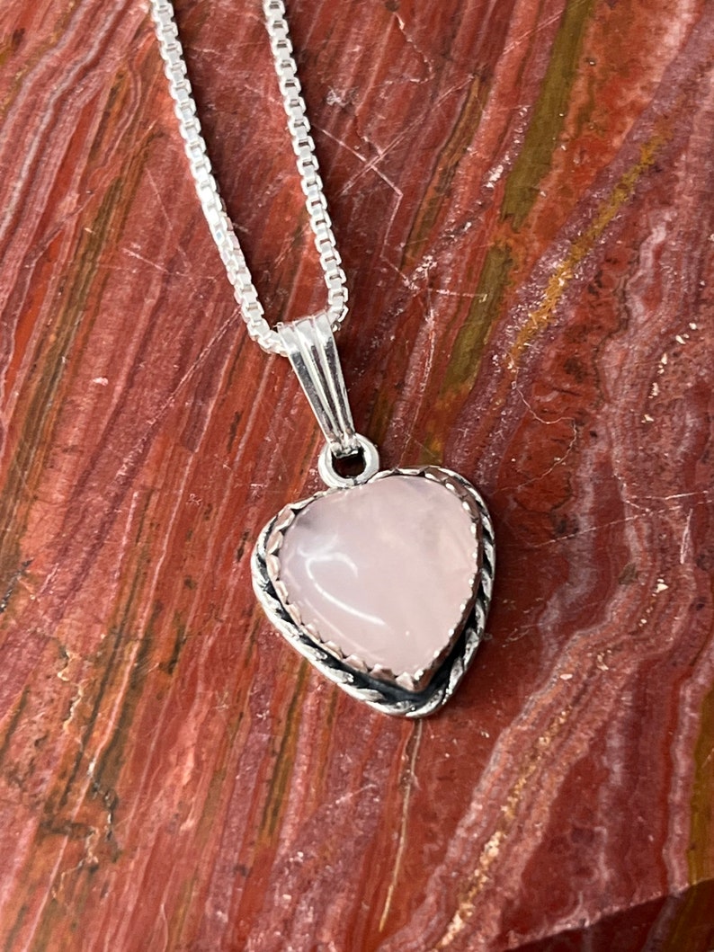 Puede incluir: Un collar de cadena de plata con un colgante en forma de coraz&oacute;n rosa. El colgante tiene un borde plateado con un dise&ntilde;o delicado.