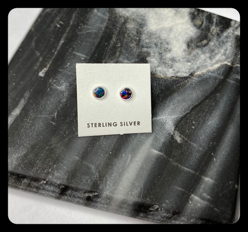 May include: A pair of sterling silver stud earrings with round, iridescent opal stones. The earrings are displayed on a white card with the text "Sterling Silver".