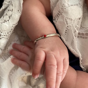 May include: A silver baby bracelet with a small, round, red stone. The bracelet is on a baby's wrist, with the baby's hand and arm visible. The background includes white lace fabric.