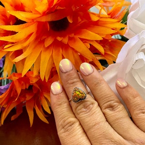 May include: A silver ring with a heart-shaped opal gemstone. The opal is a mix of orange, yellow, and green colors.