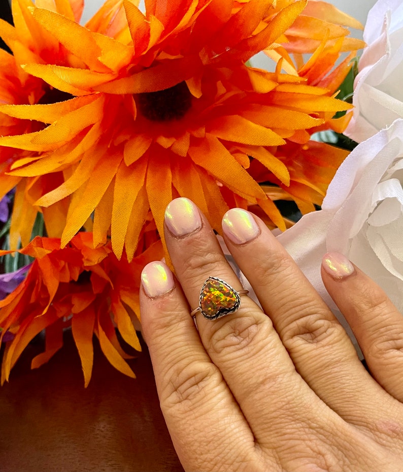 May include: A silver ring with a heart-shaped opal gemstone. The opal is a mix of orange, yellow, and green colors.