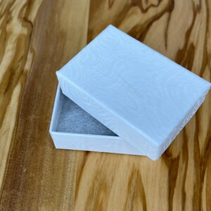 Puede incluir: Una caja de regalo rectangular blanca con un patr&oacute;n texturizado ondulado en la tapa. La caja est&aacute; abierta y el interior est&aacute; forrado con papel de seda blanco.