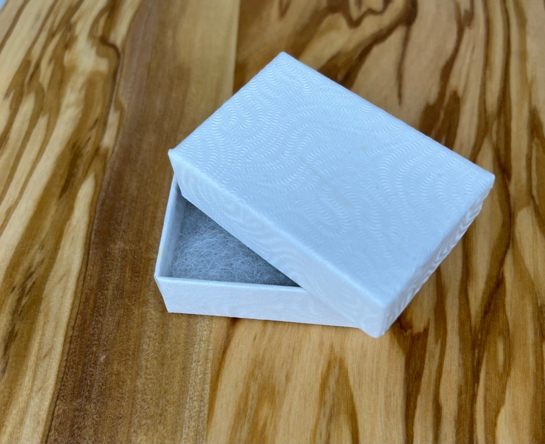Puede incluir: Una caja de regalo rectangular blanca con un patr&oacute;n texturizado ondulado en la tapa. La caja est&aacute; abierta y el interior est&aacute; forrado con papel de seda blanco.