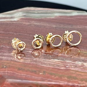 May include: Three gold hoop earrings of different sizes. The earrings are on a red and brown stone surface.