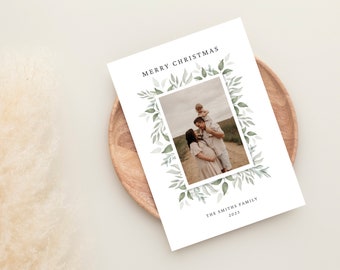 Può includere: Biglietto di Natale bianco con foto di famiglia incorniciata da un design di foglie ad acquerello. Il testo "MERRY CHRISTMAS" è in alto, con "THE SMITHS FAMILY 2023" in basso. Il biglietto è su un piatto di legno.