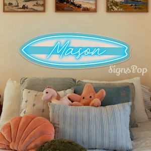 May include: A neon sign in the shape of a surfboard, with the name "Mason" in cursive script. The sign is blue and white, mounted above a bed with decorative pillows and stuffed animals. The text "SignsPop" is in the lower right corner.