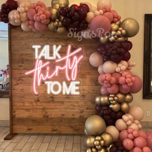 Może przedstawiać: Drewniane tło z r&oacute;żowym neonowym napisem "Talk Thirty To Me" ozdobione girlandą z bordowych, r&oacute;żowych i złotych balon&oacute;w.