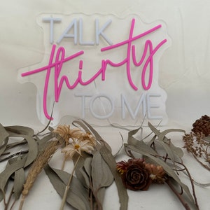 Może przedstawiać: R&oacute;żowy neonowy znak z tekstem "Talk Thirty To Me" na białym tle. Znak stoi na stole z suszonymi kwiatami i liśćmi.