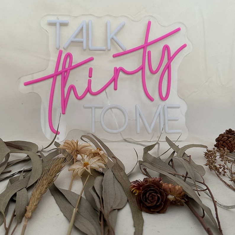 Może przedstawiać: R&oacute;żowy neonowy znak z tekstem "Talk Thirty To Me" na białym tle. Znak stoi na stole z suszonymi kwiatami i liśćmi.
