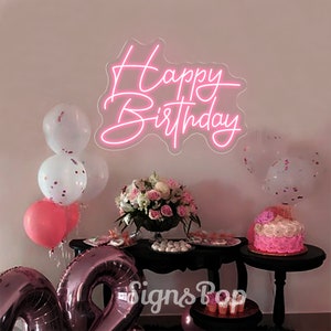 May include: Pink neon sign with the text "Happy Birthday" on a white wall. The sign is surrounded by balloons and a table with a cake and other decorations.