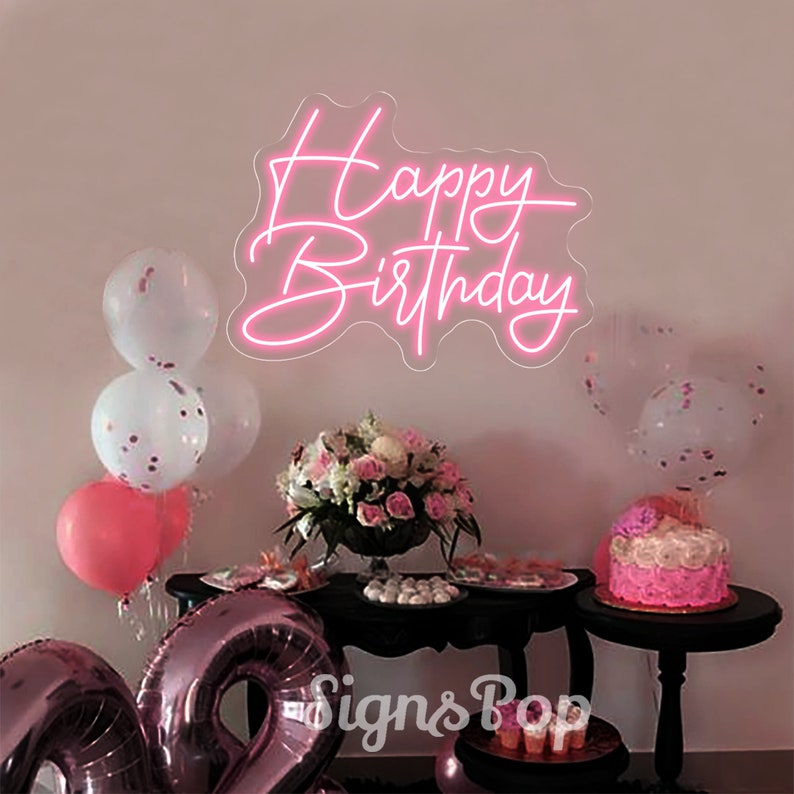 May include: Pink neon sign with the text "Happy Birthday" on a white wall. The sign is surrounded by balloons and a table with a cake and other decorations.