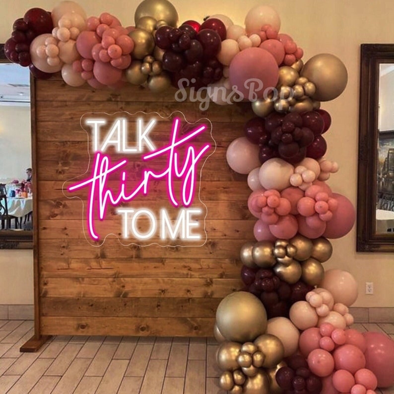Może przedstawiać: Drewniane tło ozdobione girlandą r&oacute;żowych, bordowych i złotych balon&oacute;w. Na tle znajduje się neonowy napis "Talk Thirty To Me".