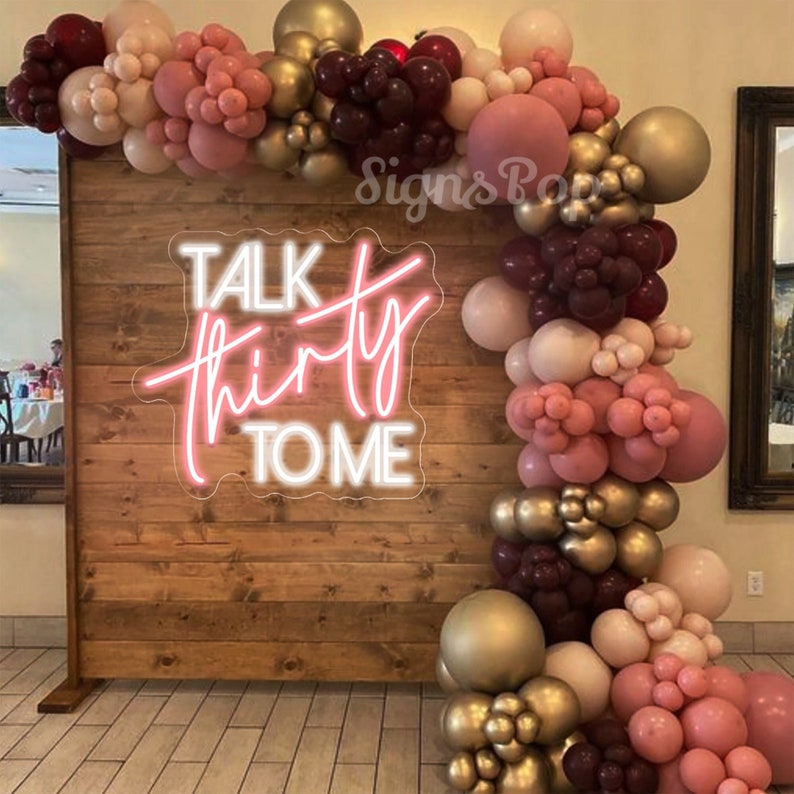 Może przedstawiać: Drewniane tło z r&oacute;żowym neonowym napisem "Talk Thirty To Me" ozdobione girlandą z bordowych, r&oacute;żowych i złotych balon&oacute;w.