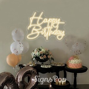 May include: A white neon sign with the words "Happy Birthday" on a white wall. The sign is surrounded by balloons and a table with a cake and other decorations.