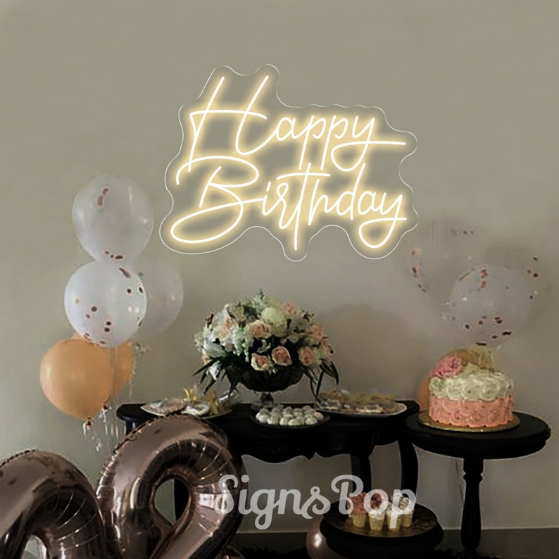May include: A white neon sign with the words "Happy Birthday" on a white wall. The sign is surrounded by balloons and a table with a cake and other decorations.