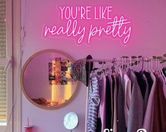May include: Pink neon sign with the text "You're like really pretty" on a pink wall with a round mirror and a clothing rack with clothes hanging on it. The text "SignsPop" is visible in the bottom right corner of the image.
