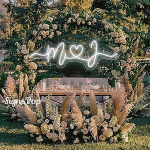 May include: A white neon sign with the letters "M" and "J" connected by a heart. The sign is hanging in front of a floral archway with pampas grass and white flowers. The sign says "M♥J".