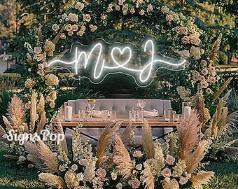 May include: A white neon sign with the letters "M" and "J" connected by a heart. The sign is hanging in front of a floral archway with pampas grass and white flowers. The sign says "M♥J".