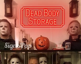 May include: A red neon sign with the words "DEAD BODY STORAGE" in a decorative frame. The sign is surrounded by Halloween-themed decorations, including a carved pumpkin, Michael Myers masks, and miniature figures. Two lanterns hang on the wall.