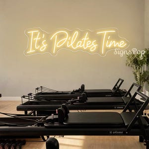 May include: A Pilates studio with black Pilates reformer machines. A neon sign in a cursive font above the machines reads "It's Pilates Time". The sign is a warm yellow colour. The studio has a clean, minimalist aesthetic, with a focus on the equipment.