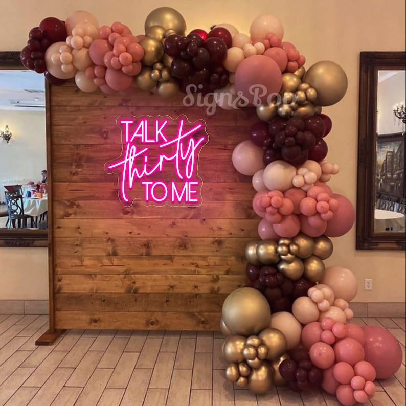 Może przedstawiać: Drewniane tło z r&oacute;żowym neonowym napisem "Talk Thirty To Me" ozdobione girlandą balon&oacute;w w odcieniach r&oacute;żowego, bordowego i złotego.